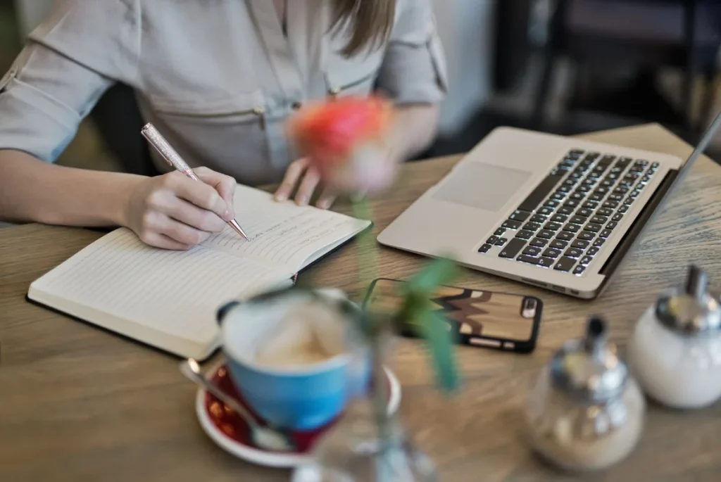 Menulis catatan di buku dengan laptop dan kopi di meja